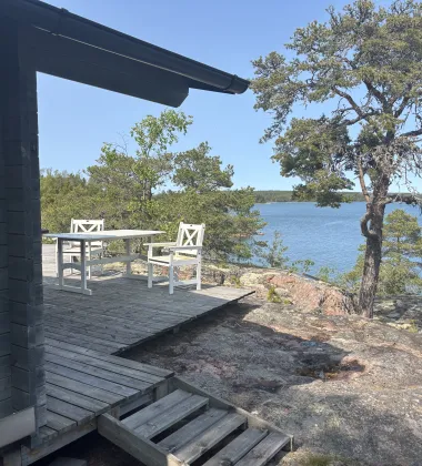 En stuga med en altan ut mot havet med två vita trästolar och en stor vindpinad tall. 