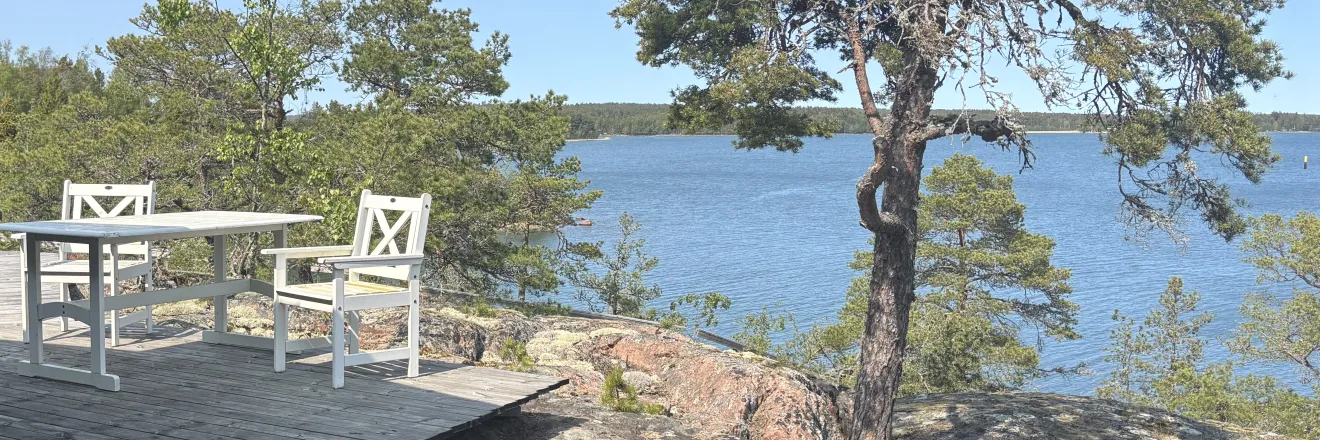 En stuga med en altan ut mot havet med två vita trästolar och en stor vindpinad tall. 
